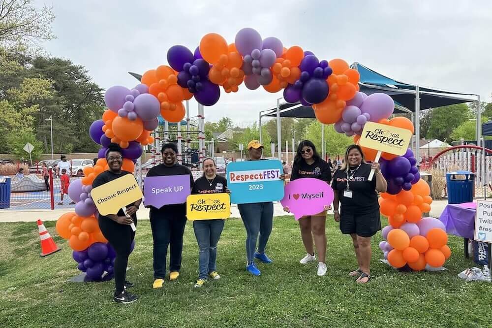 A group of six adults underneath an arch of orange and purple balloons holding up signs relating to dating violence and prevention