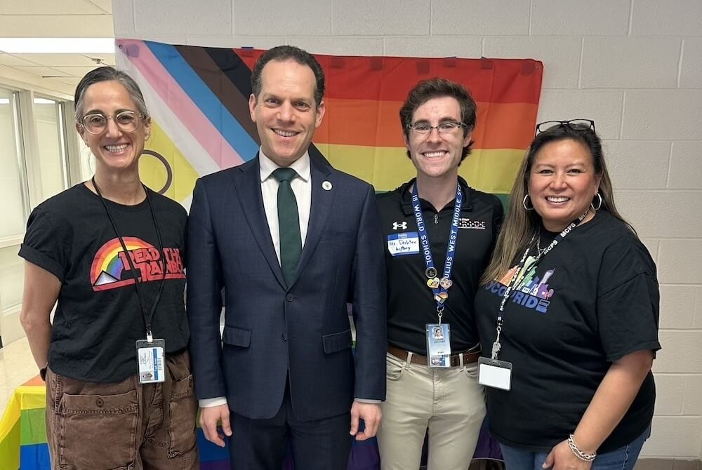A group of MCPS Staff alongside Councilmember Evan Glass stand smiling in front of a rainbow flag