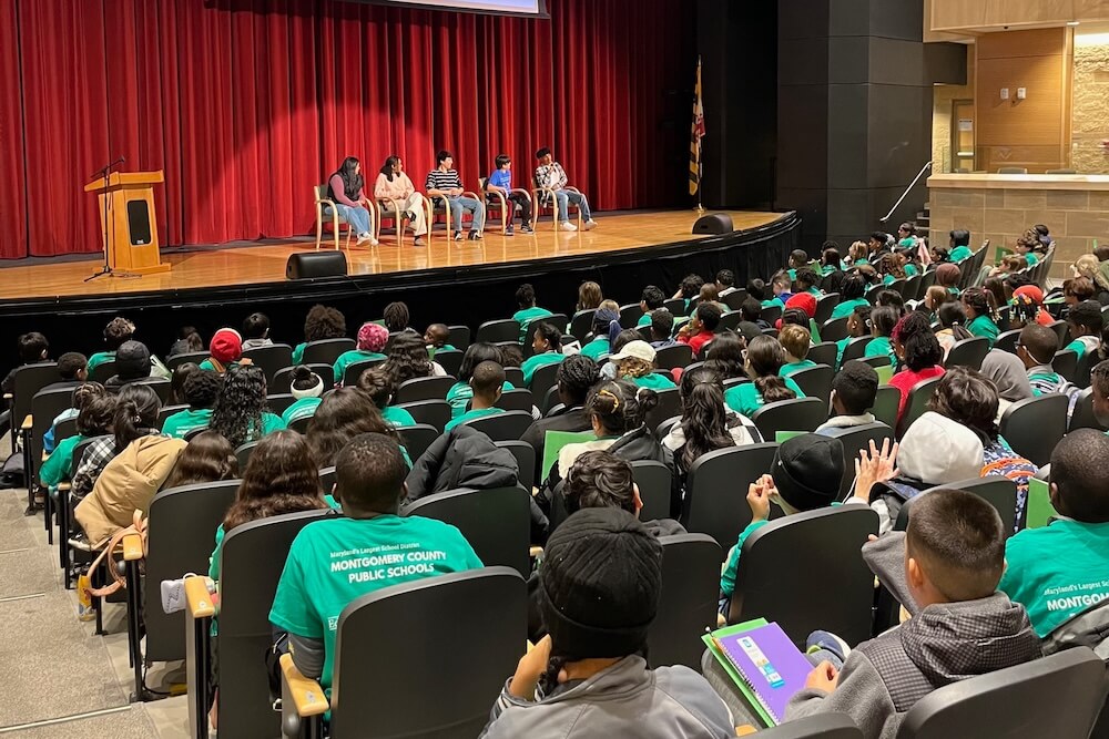A group of elementary school students in green t-shirts sitting in an auditorium listening to five speakers on a stage